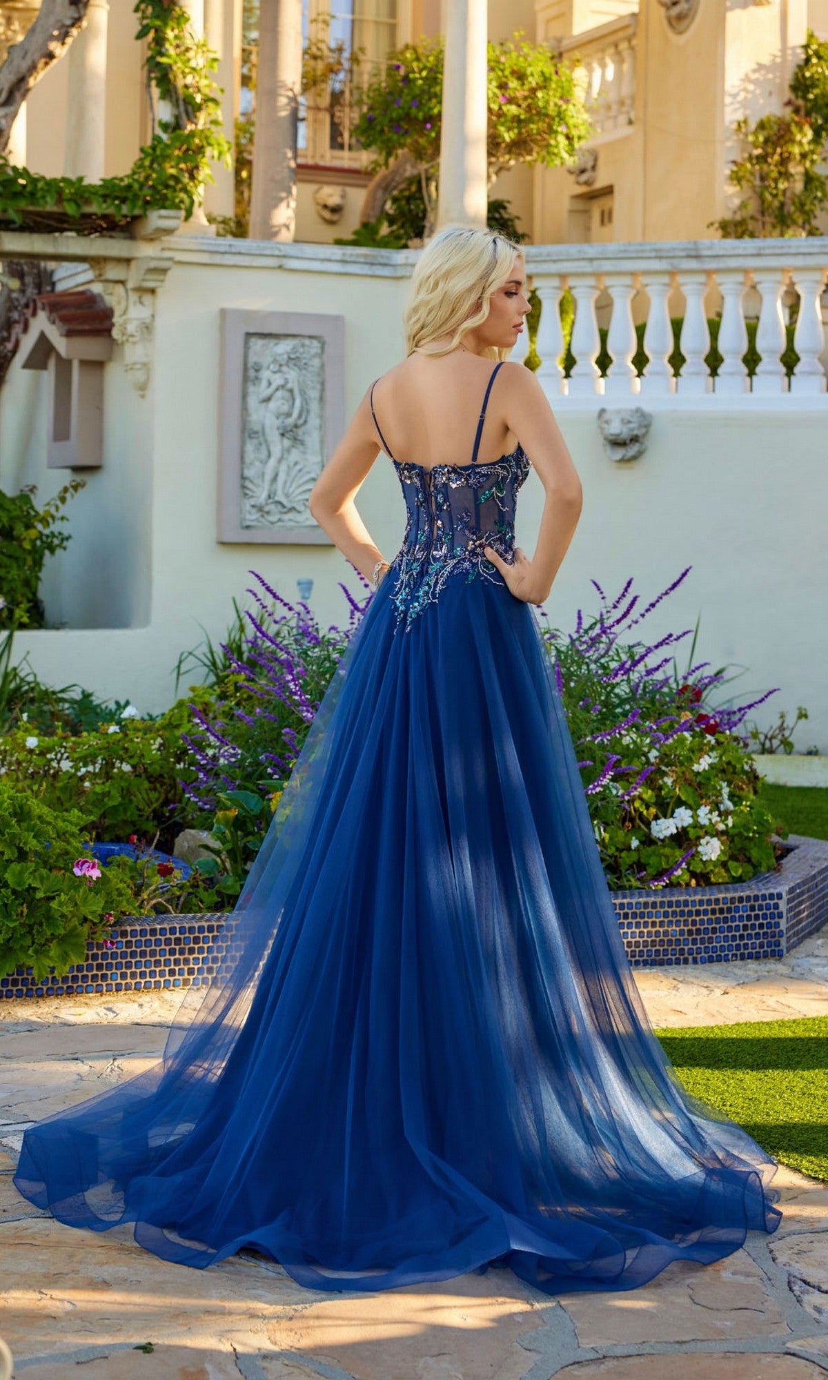 A woman wearing the Juliet JT2640G long blue tulle prom dress with beaded details and a lace-up back stands on a stone patio outdoors, facing away, with flowers and a large building in the background.